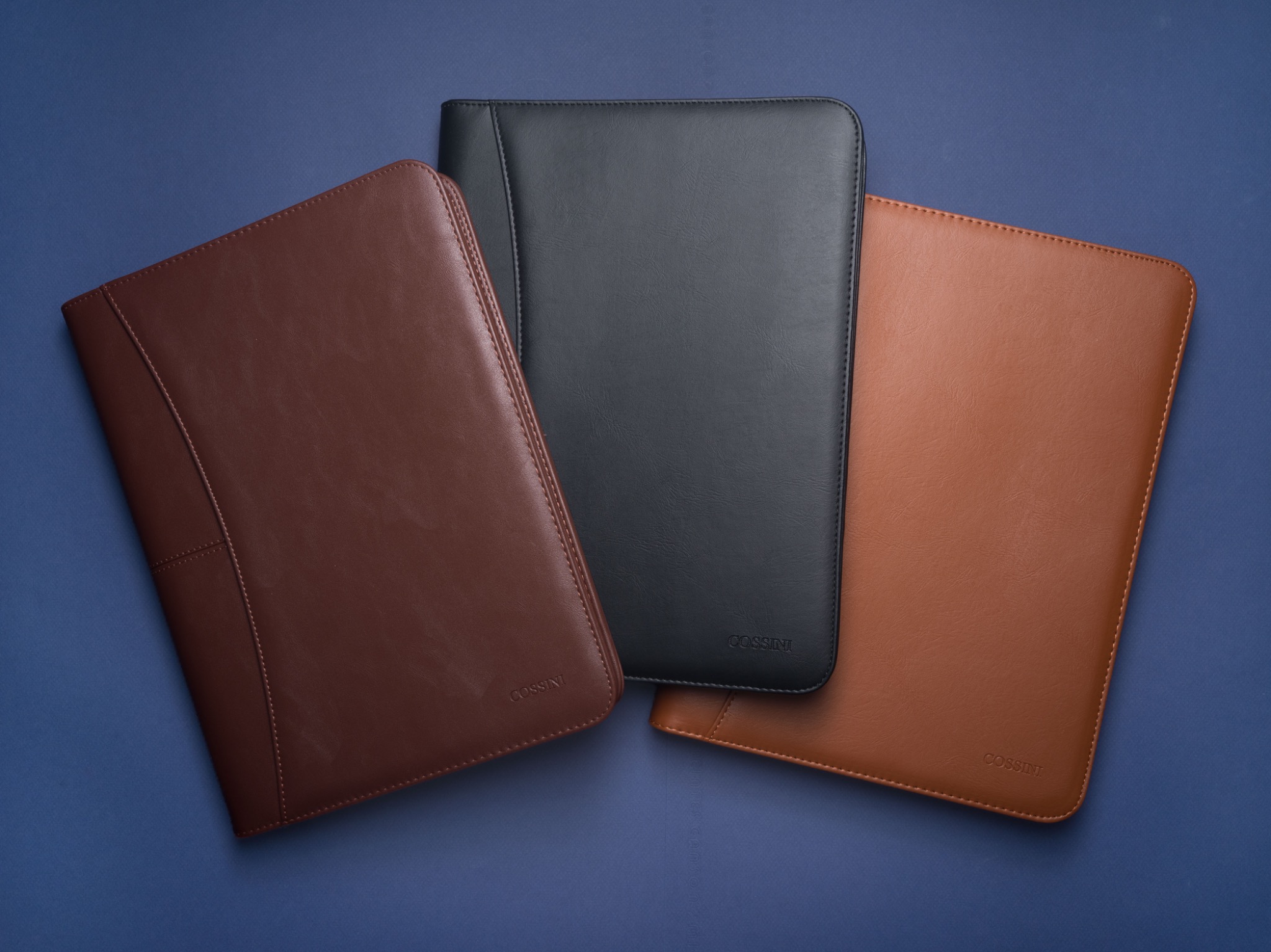 Three Cossini leather padfolios arranged on a deep blue surface